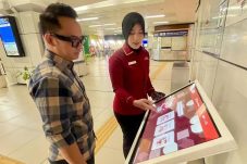 Petugas LRT Jabodebek menjelaskan penggunaan layar interaktif kepada seorang penumpang di area stasiun untuk layanan survei kepuasan dan pengaduan digital real-time.