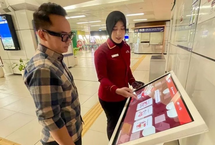 Petugas LRT Jabodebek menjelaskan penggunaan layar interaktif kepada seorang penumpang di area stasiun untuk layanan survei kepuasan dan pengaduan digital real-time.